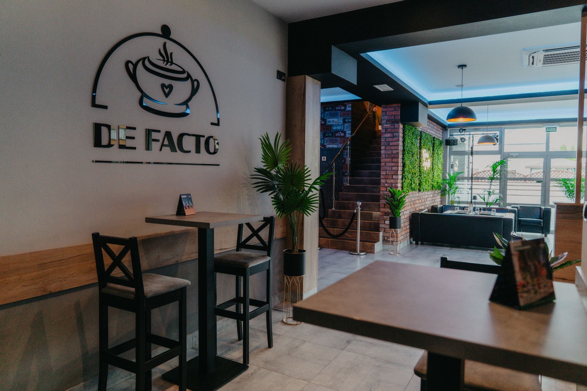 Wnętrze restauracji De Facto z logo na ścianie, wysokie stoliki i zielona ściana